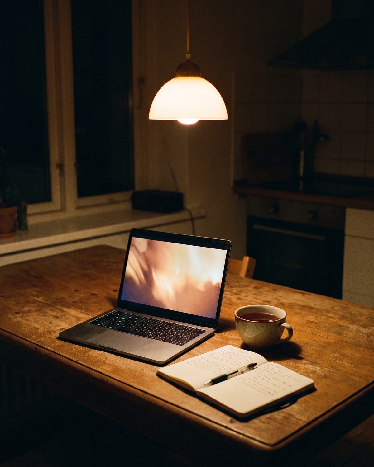An open laptop on a kitchen table at night, a lamp on, a cup of tea and a notebook